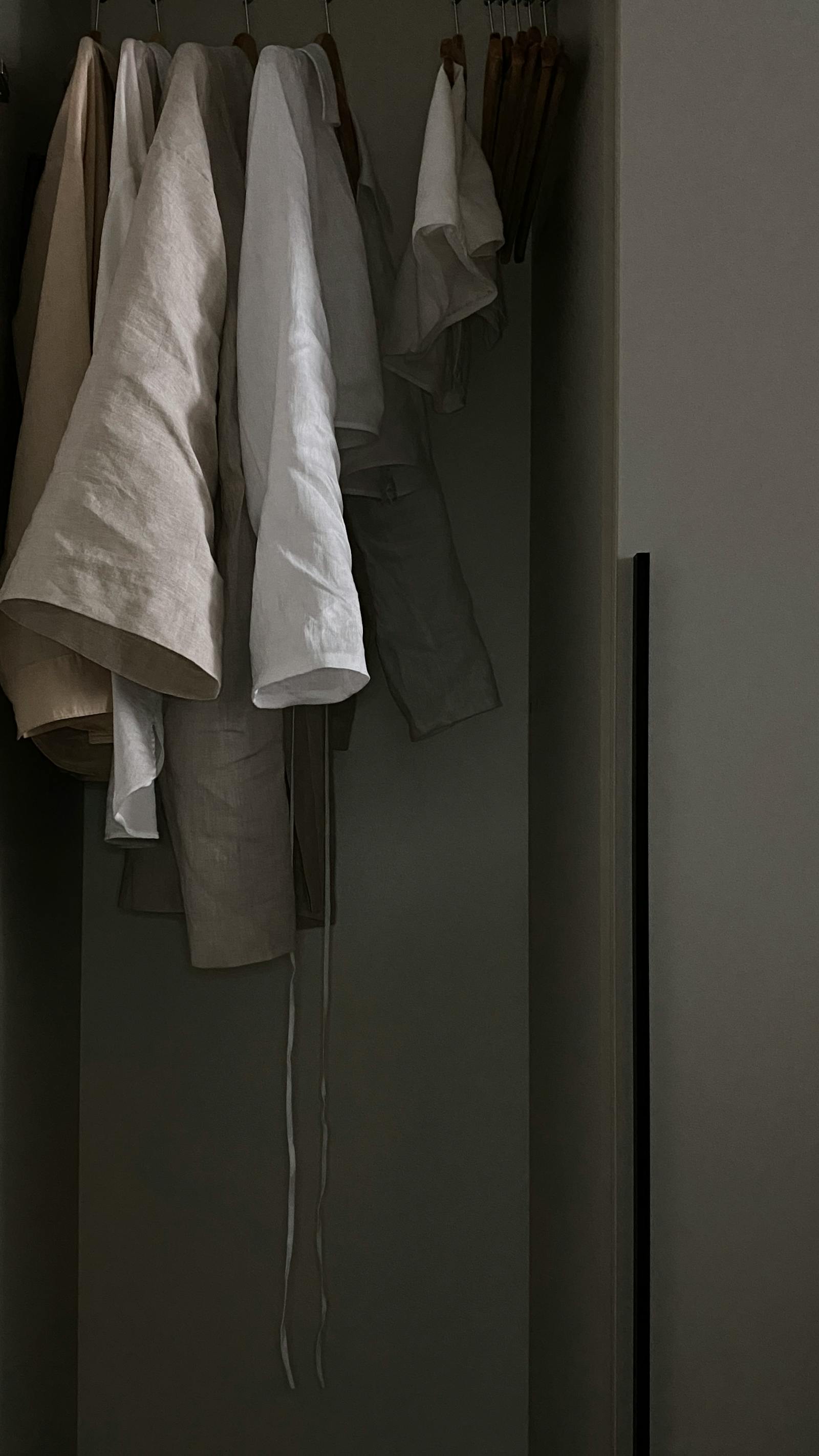 Cream and oat-coloured linen pieces hanging on wooden hangers in a dim closet